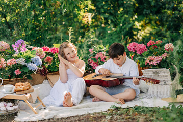 Kid boy playing guitar sitting on the grass in the park with summer flowers on picnic with girl. Children romantic friends lovely
