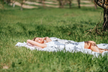 woman sleeps on a white bed in the fresh spring grass in the garden. Dressed in a blue nightgown.