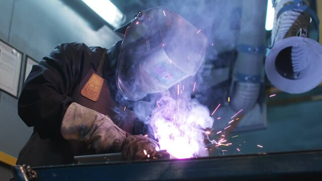 Asian man is focused on work. Uniformed welder lifts protective mask and examines detail. Male is satisfied with result and smiles. Men's labor. Diversity of men's profession.
