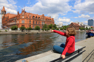 A girl sits on a parapet on the banks of the Brda River in Bydgoszcz. People visit riverfront in Bydgoszcz. Bydgoszcz is the capital of region Kujawsko-Pomorskie visited by 2.25m tourists annually  - obrazy, fototapety, plakaty
