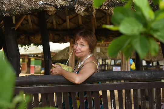 Attractive woman sitting at the tropical alcove and drinking coconut juce on the beach. Senior woman wear the dress. Concept of active retired elderly and travel lifestyle.