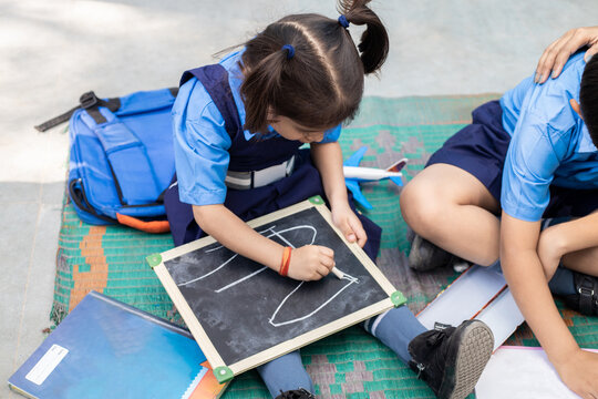 Cute little indian girl student wearing school uniform writing on slate board with chalk while sitting on floor. Government school. Rural india. Education concept. Rural india. Top view.
