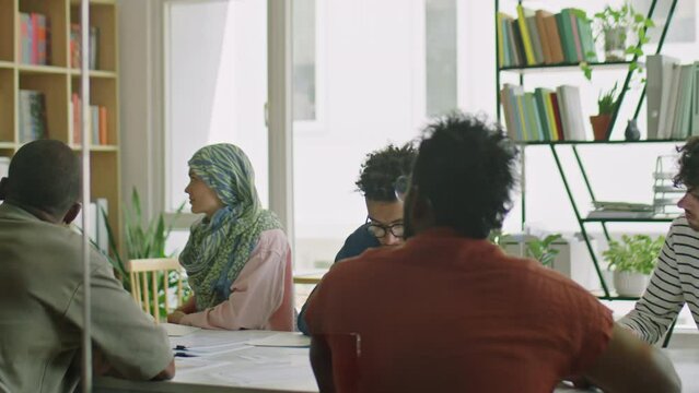 Group of muti-ethnic immigrant students listening to female teacher as she standing by whiteboard and showing educational cards during lesson in classroom