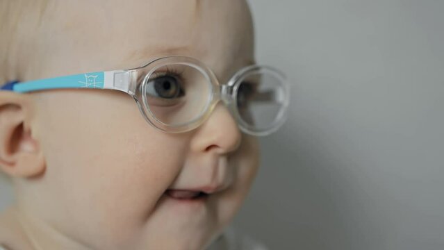 One year old baby with glasses opens mouth and sticks out tongue. Caucasian infant is watching TV at home. White kid has poor eyesight or bad vision. Small child has eye disease, squint or strabismus
