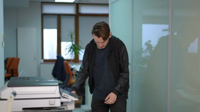 Young man checking broken printer in office thinking. Side view Caucasian male employee with equipment indoors at workplace
