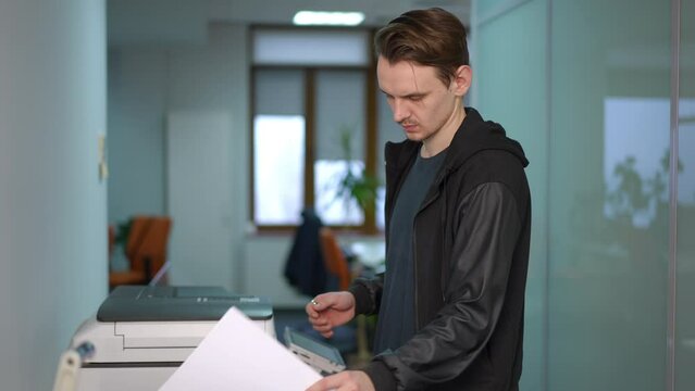 Side view portrait of troubled unsure young man with paperwork at printer in office. Caucasian male employee hesitating photocopying documents at workplace indoors