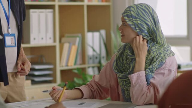 Female teacher helping muslim girl in hijab with doing assignment in classroom during lesson