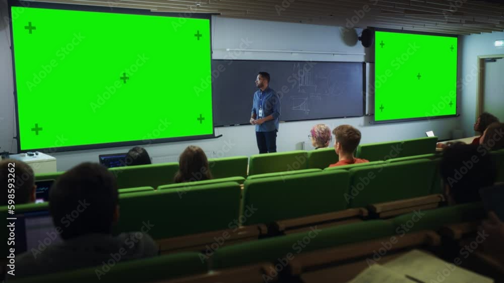 Poster Young University Professor Giving a Lecture, Pointing on a Green Screen Mock Up Display. Group of Talented Diverse Students Study in Modern Classroom. Teacher Showing Content on Two Big Screens