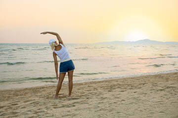 Fototapeta na wymiar older woman wearing sportswear doing side stretching exercises on seaside in summer day at sunset.