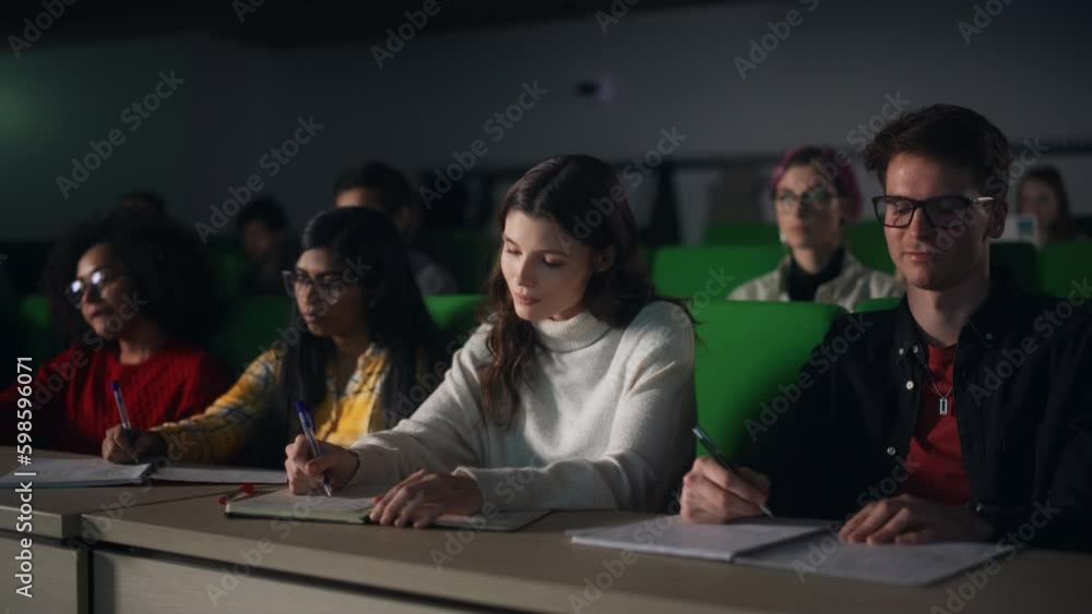 Poster Thoughtful Beautiful Young Female Studying in University, Listening to a Lecture in a Crowded Room with Diverse Multiethnic Classmates. Students Listening to Lecture and Taking Notes in Notebook