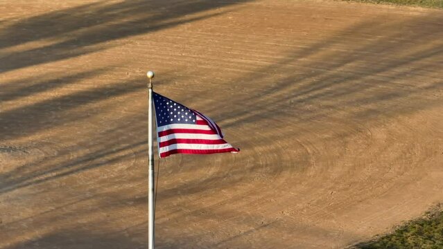 American Flag Waving With Baseball Field Dirt Infield In Background. Establishing Shot Of USA Past Time, Baseball. Little League Field. Aerial View.