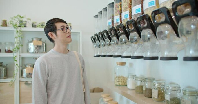 Man with reusable shopping bag buying eco bio organic vegan products in zero waste shop. Asian male in plastic free grocery store. Small business startup go green market for reduce climate change.