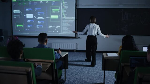 Young Teacher Giving a Web Development Presentation to a Diverse Multiethnic Group of Female and Male Students in a Modern College Room. Female Walking Between the Rows While Giving a Lecture