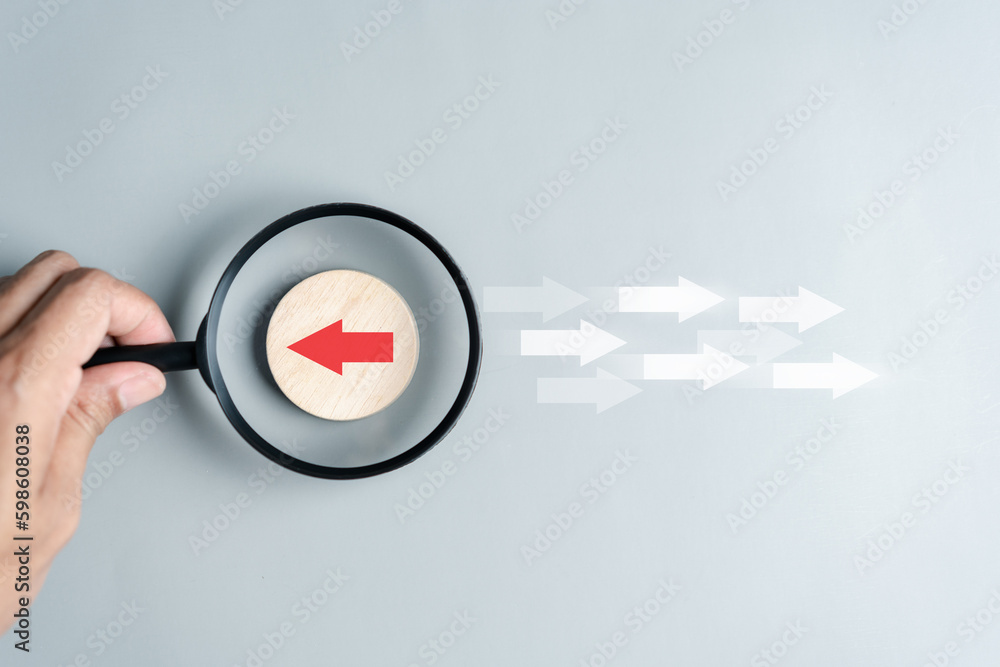 Fototapete Hand holding magnifier focus to Red arrow and different direction facing opposite direction with white arrow for business disruption and technology transformation and different thinking idea concept.