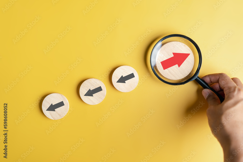 Leinwandbilder Hand holding magnifier focus to Red arrow and different direction facing opposite direction with black arrow for business disruption and technology transformation and different thinking idea concept.