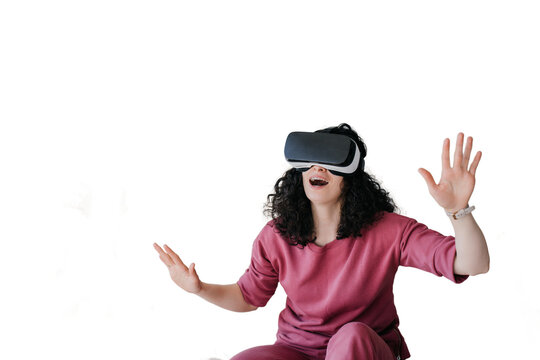 Amazed curly caucasian young woman in lilac clothes sitting on bed spreading hands using vr glasses at bedroom. Happy girl in virtual reality headset having fun against transparent background.