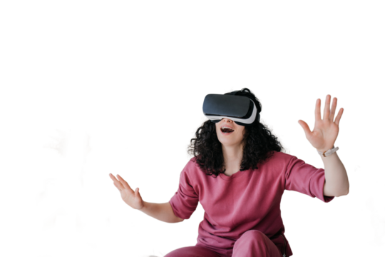 Amazed curly caucasian young woman in lilac clothes sitting on bed spreading hands using vr glasses at bedroom. Happy girl in virtual reality headset having fun against transparent background.