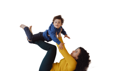 Happy Italian mother older sister lying on back against transparent background having fun with little kid lifting excited child boy in air on outstretched legs. Curly mom play airplane with son
