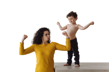 Sporty caucasian curly woman at kitchen with son demonstrating her biceps and curly son standing on table showing biceps. Italian young babysitter spending time with kid against transparent background