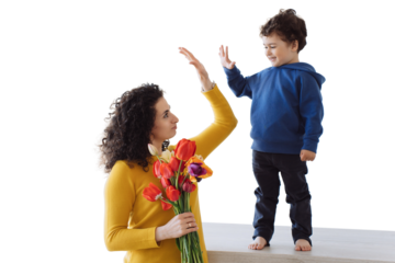 Cheerful young woman in sweater holding colourful flowers giving five to her little son standing on table, smiling. Little curly kid  congratulates mom on mother's day against transparent background