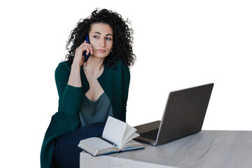 Exhausted  young curly woman in green cardigan sitting at desk with laptop and notebook making call by phone looking aside. Calm Spanish entrepreneur remote working against transparent background.