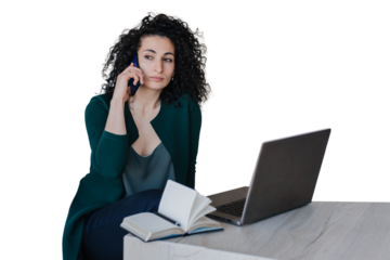Exhausted  young curly woman in green cardigan sitting at desk with laptop and notebook making call by phone looking aside. Calm Spanish entrepreneur remote working against transparent background.