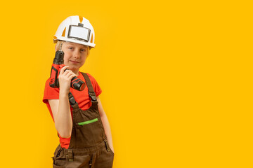 Child wears helmet and working overalls holds drill in his hands. Boy 9-10 years old imitates builder, yellow background. Copy space