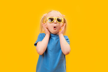 Preschool girl in blue T-shirt on yellow background wears glasses and makes surprised face. Studio portrait.