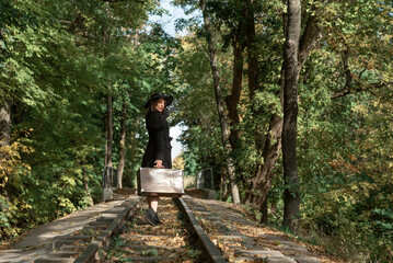 Woman leaves with a suitcase along the rails through the autumn forest. Woman in black clothes. Back view