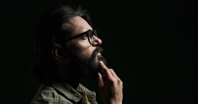 Closeup portrait of serious young man touching beard and looking away while contemplating against black background Generative AI