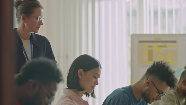 Female teacher walking around diverse migrant students and talking to them during exam in language school