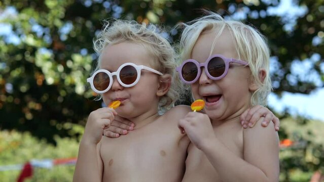 Low angle cinematic shot of two caucasian naked twin sisters hugging, licking candy lollipops. Girls in cool glasses in nature in park eat lollipops in sunny weather on weekend. Day off Generative AI