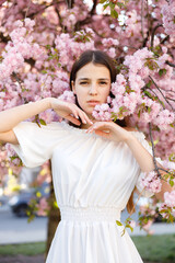 Cute Caucasian Dark Haired Girl Standing in the Park, Posing with Hands Near Sakura Tree With Pink Flowering. Femininity and Natural Beauty Concept
