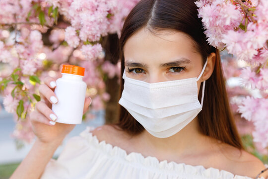 Portrait of Young Caucasian Woman Wearing Protective Mask with Unhealthy Look Holding Little Bottle of Medicines in Blossoming Park. Covid During Spring Season