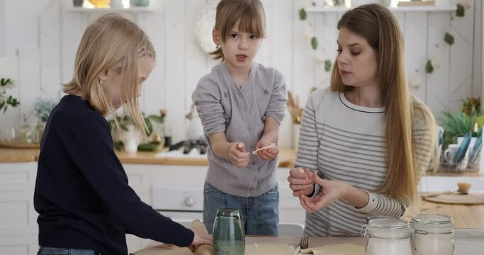 Young mother rolling dough and guiding cute daughters in making fresh biscuits on kitchen table at home Generative AI