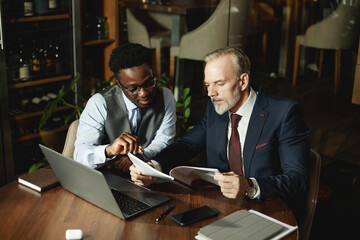 Fototapeta premium Colleagues examining contract together while sitting at table with laptop during their meeting in cafe