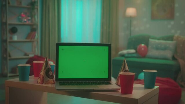 Laptop with green screen display in front of a room decorated for a house party. On a blurred background, lights flash and a disco ball sparkles. Advertising area, workspace mock up.