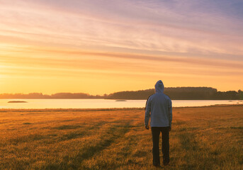 Young caucasian man in hoodie walking to pond under dramatic sunrise sky. Czech landscape