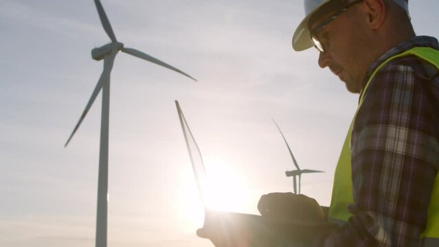 Technician engineer in a white hard hat and overalls at a wind farm checks the condition of the turbines using a laptop. Professional technician wearing grey overalls, orange helmet and Generative AI