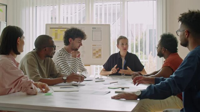 Group of multi-ethnic immigrant students sitting around table in classroom and listening to female teacher during lesson
