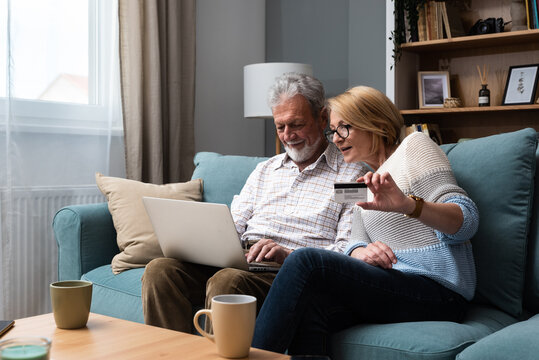 Senior Couple At Home. Handsome Older Man And Attractive Older Woman Are Spending Time Together. Sitting On A Sofa With Laptop Computer And Credit Card In Hands. Online Shopping Ordering Or Reserving