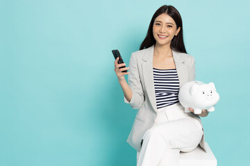 Portrait of Happy Asian businesswoman holding mobile phone and white piggy bank isolated on green...