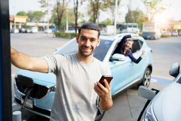 Happy handsome Asian male driver preparing to recharge his electric vehicle or EV car at EV fast charge station during the trip. Future and sustainable transportation concept.