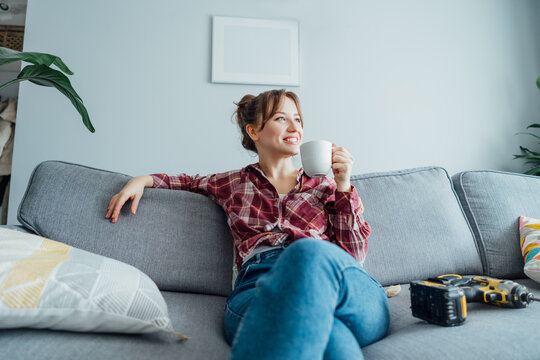 Happy smiling proud of herself young woman relaxing with coffee cup after she puts by her own a picture on the wall at her home. Housekeeping work. Doing repair herself. DIY, gender equality concept