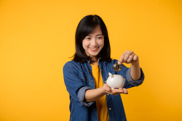 Happy young asian woman wearing yellow t-shirt denim shirt saving digital crypto currency to piggy bank isolated on yellow background. Digital money saving investment concept.