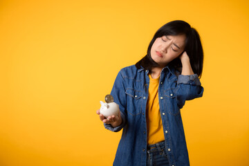 Unhappy asian young woman wearing yellow t-shirt denim shirt pulling digital coin crypto currency out of piggy bank isolated on yellow background. Payment digital money debt financial concept.