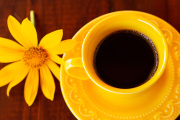 Uma xícara amarela com café sobre píres e um girassol, em cima de uma mesa de madeira escura.