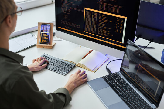 Rear view of young programmer writing security codes on computer connecting laptop and smartphone sitting at his workplace