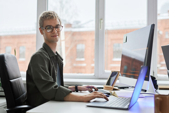 Portrait of young programmer in eyeglasses looking at camera while developing software on computer at his workplace - Powered by Adobe