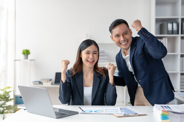 Two young Asian business people successful excited raised hands rejoicing with a laptop computer in a modern office.